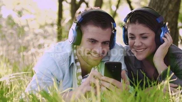 Couple listening to music/ selecting songs on smart-phone in summer on headphones while hanging out in park.