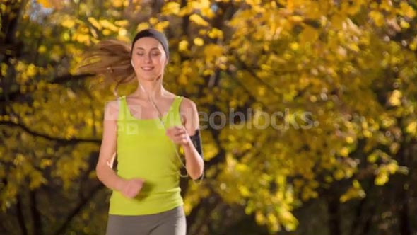Running woman. Runner is jogging in sunny bright light in the autumn park background. Female fitness model training outside on a warm fall day and listening to music. Sport lifestyle. 4k slow motion.